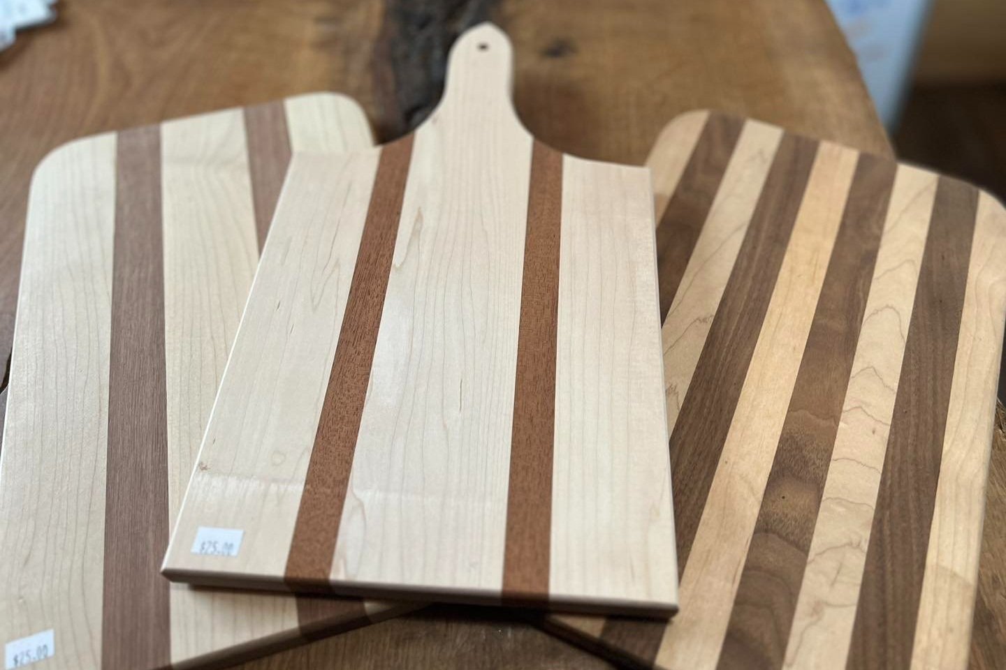 432951290_835510395267454_9024665499014404090_n Three wooden cutting boards with alternating light and dark wood stripes displayed on a rustic wooden table. The boards feature a mix of maple and cherry wood, showcasing craftsmanship and design.