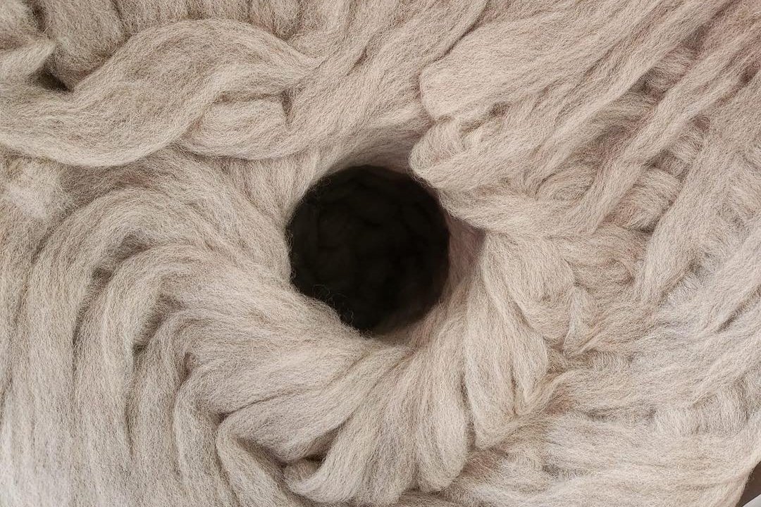 433647142_826314172853743_8578758367437446271_n Close-up of a soft, light beige wool yarn ball featuring a central hole, showcasing the texture and fiber details ideal for knitting and crafting projects.