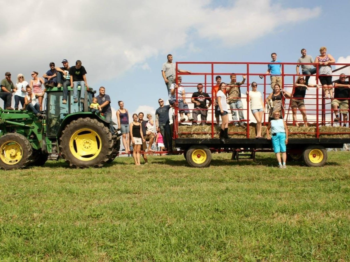 Emily+Kalenauskas+-+352069554_794542055354219_5043396710749093059_n Group of people enjoying a farm day, posing on a green tractor and a red hay wagon in a sunny field, surrounded by grassy landscape and blue skies.