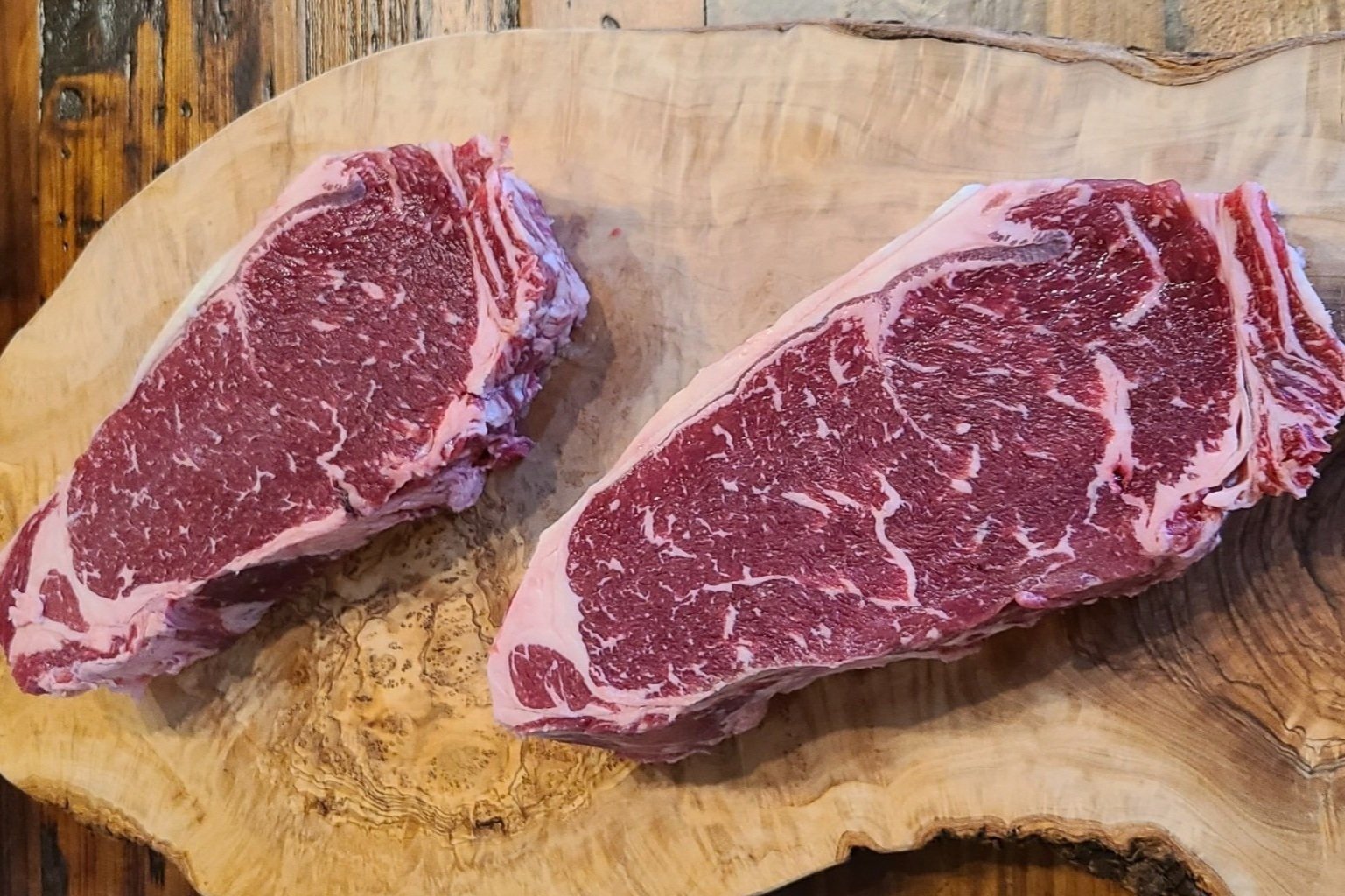 emily+-+123_1 Two marbled ribeye steaks displayed on a wooden cutting board, showcasing rich red meat and creamy white fat, ideal for grilling or pan-searing.