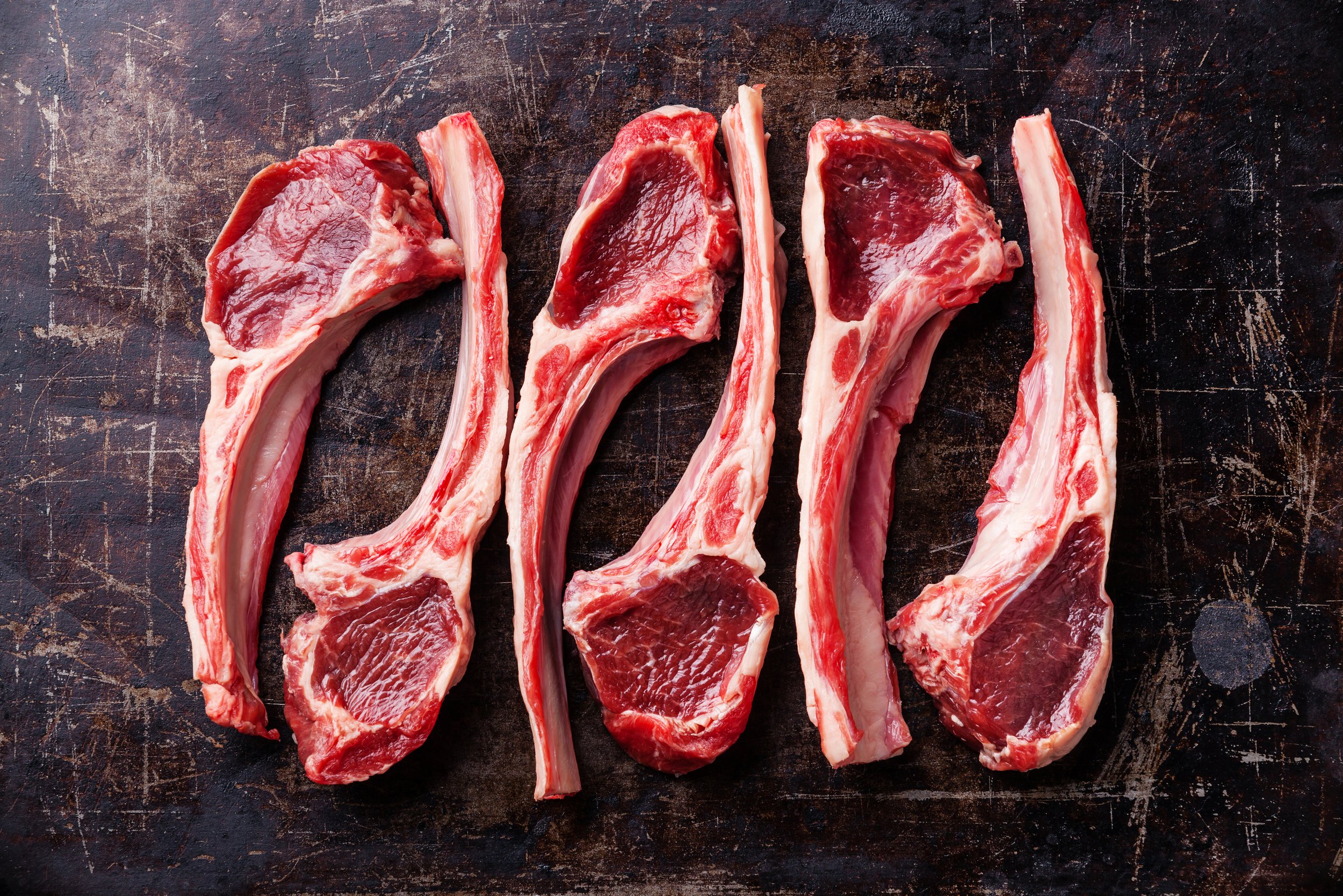 Raw fresh lamb ribs on dark background Four raw lamb chops with visible marbling, arranged on a rustic wooden cutting board, showcasing their fresh red meat and bone structure. Perfect for grilling or roasting recipes.